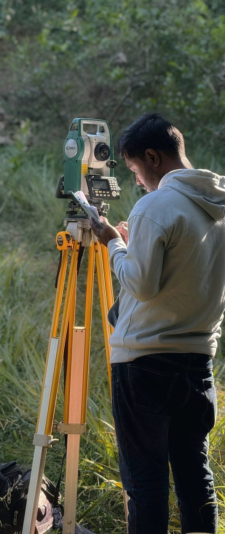 A surveyor reading a total station on a tripod in tall grass